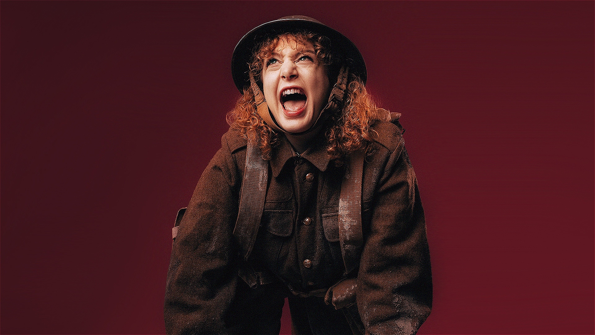 Jessica Durand is dressed in a historical World War 1 soldier's uniform covered in dirt. She leans forward with her hands, shouting with an intense expression. She has curly red hair partially covered by a helmet. The background is a deep burgundy colour