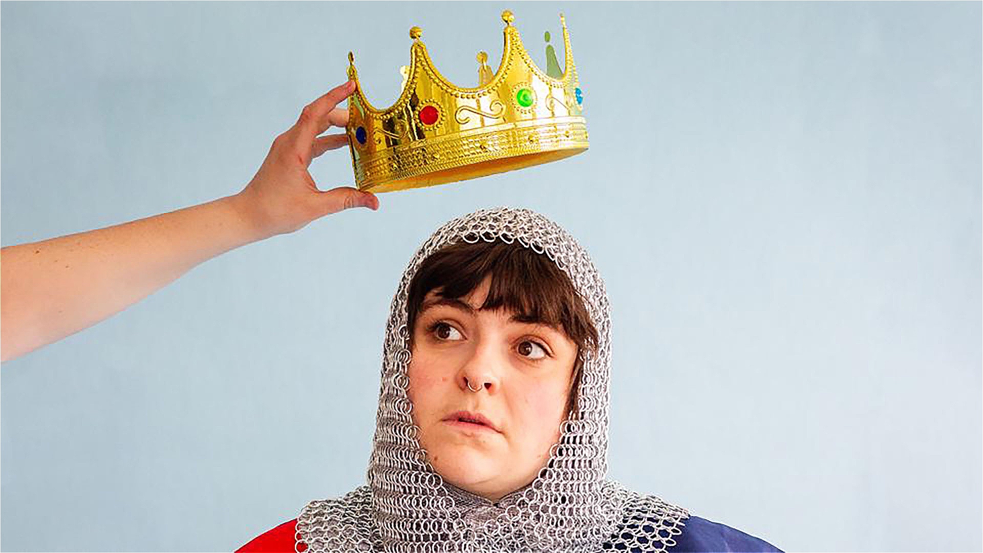 A person dressed in chainmail in front of a light blue background. They are looking off in the distance as a crown is placed on their head from above.