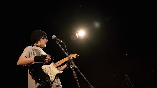 A woman dressed as an emo teenage boy stands at a mic, singing and playing an electric guitar.
