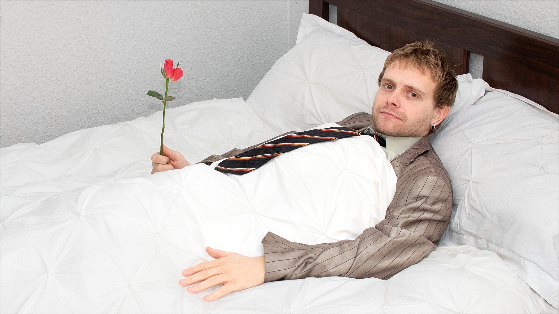 Theo Mason Wood lies in a neatly made bed, wearing a striped suit and loosened tie. He gazes directly at the camera with a subtle smirk, holding a single red rose in one hand. The crisp white bedding contrasts with his slightly disheveled outfit, creatin